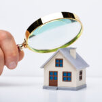 Person holding a magnifying glass over a tiny wooden home to show home inspecting.