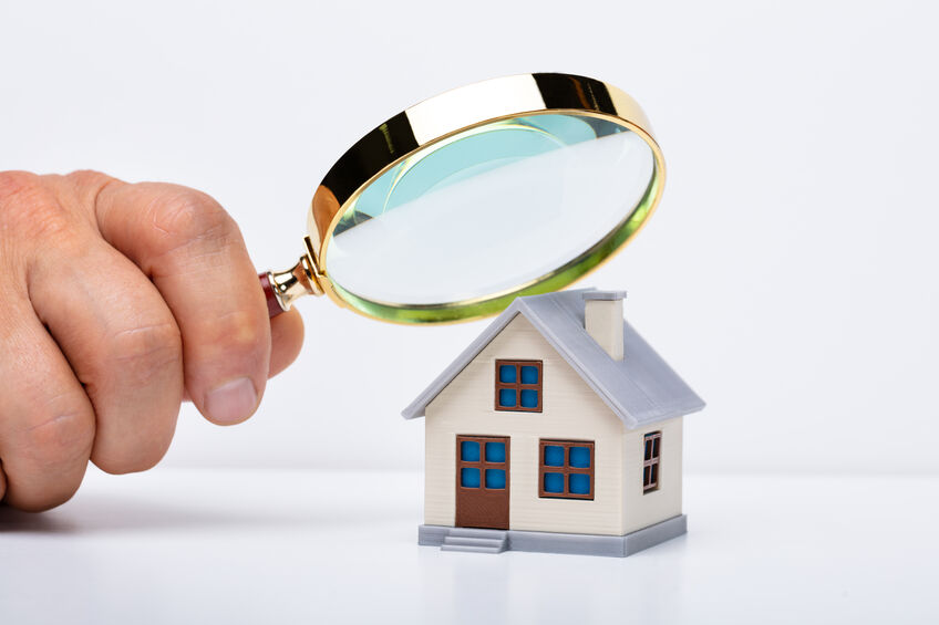 Person holding a magnifying glass over a tiny wooden home to show home inspecting.